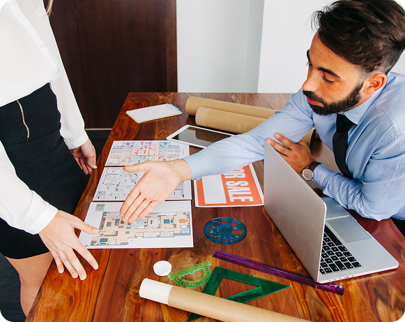 Man in a suit reviews documents while a woman with head in hand looks distressed. A laptop, coffee cup, folders, and a box are on the table. Bright room.